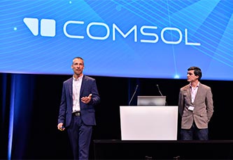 两位演讲者在台上演讲，他们之间有一个讲台，他们身后的幻灯片显示了 COMSOL 徽标。