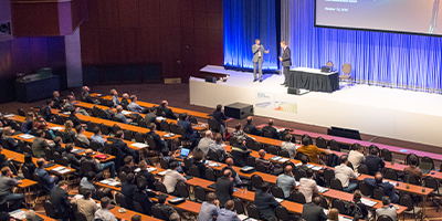 A bird's-eye view of many COMSOL Conference attendees sitting at desks during a keynote presentation, with two people presenting onstage.