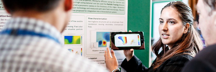 A COMSOL Conference attendee standing in front of a poster, showing two other attendees an image of a simulation on a phone-like device.