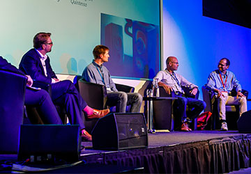 Five speakers sitting spaced apart onstage during a panel discussion; a slide containing an acoustics simulation is visible behind the speakers.