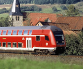 A red double-decker coach train driving through the verdant German countryside, passing a church and other rustic buildings.