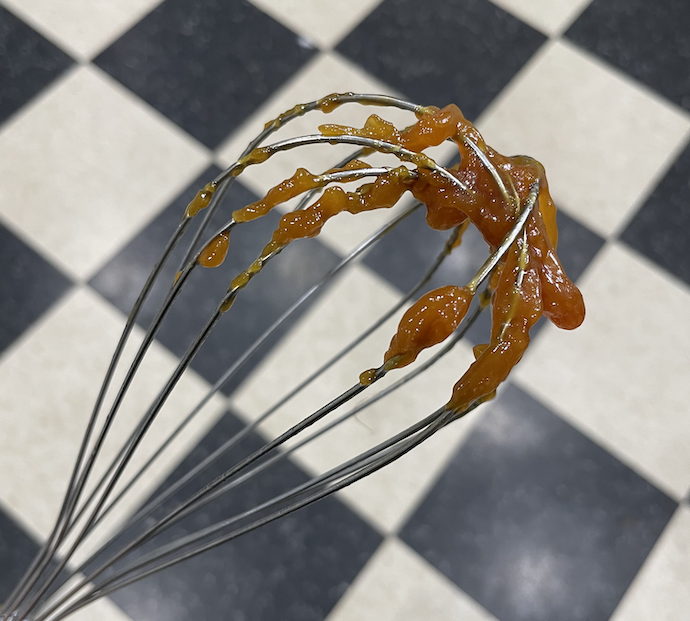 A photograph of a metal whisk with clumps of caramel on the end.