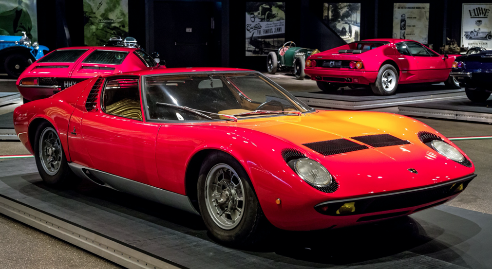 A photograph of a red Lamborghini Miura in front, with other sports cars and supercars in the background.
