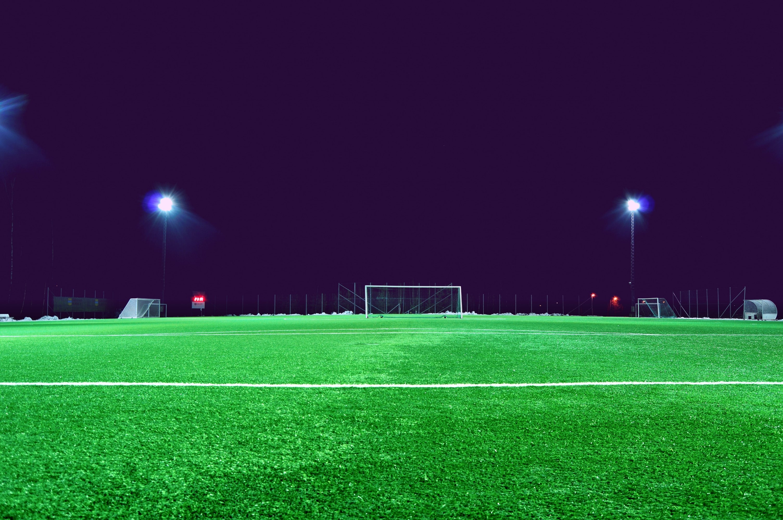 A photograph of a green soccer field at night, with a dark sky and two tall LED lights on either side of the goal post.