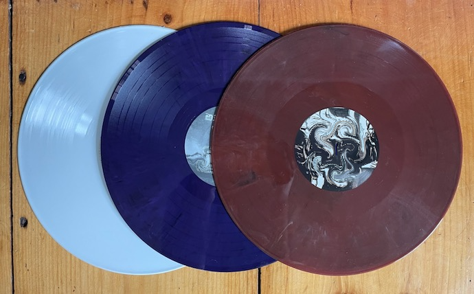 A photograph of three different-colored vinyl records resting on top of each other, with white on the left, purple in the center, and red on the right.