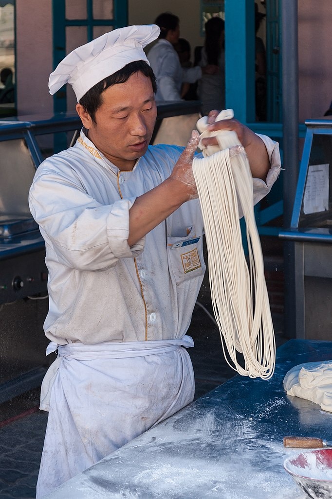 A man working at an outside counter as he hand-pulls a loop of pasta noodles.