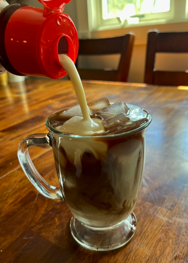 An image of oat milk being poured into a glass mug filled with iced coffee.