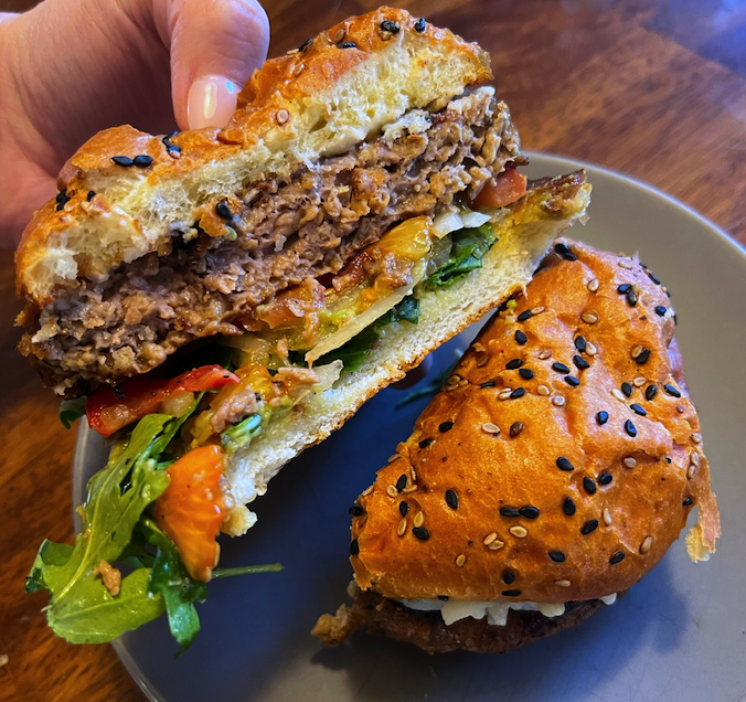 A close-up image of a plant-based burger, which consists of vegan mozzarella, crushed avocado, arugula, onion, tomatoes, pickles, a sesame seed bun, and a meat substitute.