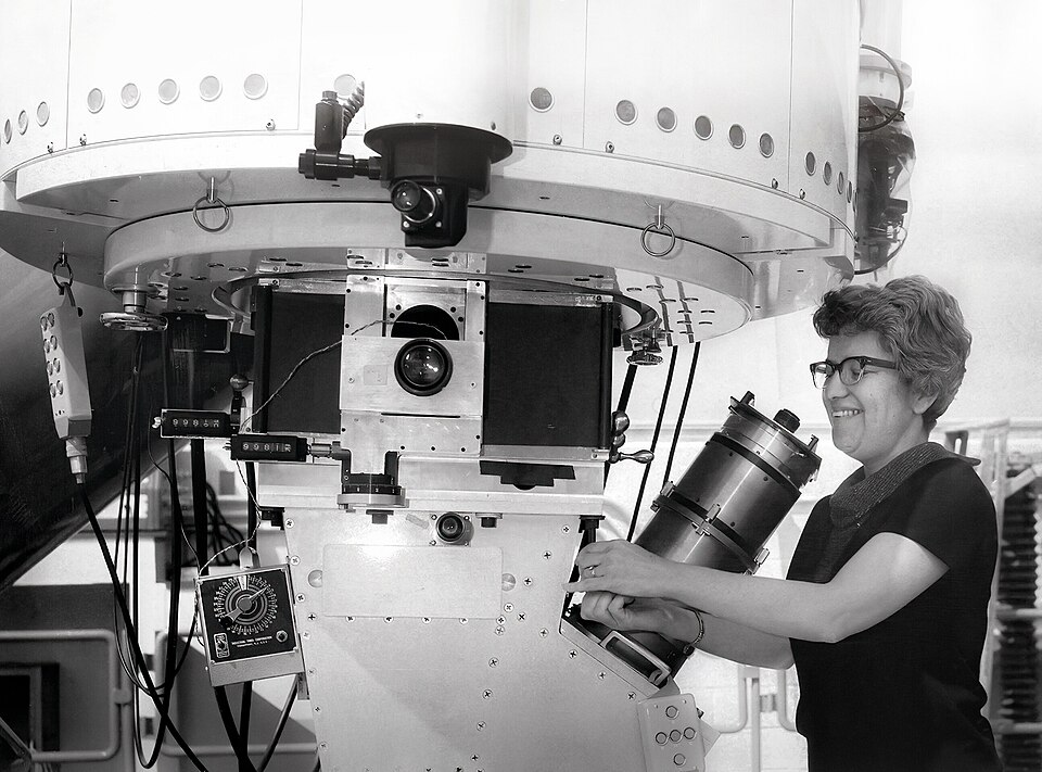 A black and white photograph of Vera Rubin using a large telescope with a spectrograph attached