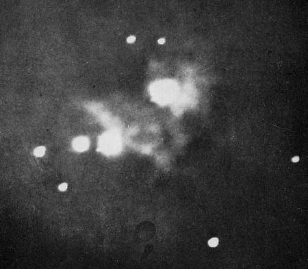 A black and white photograph of the Orion Nebula, where most of the image is dark and there is a blurry white clump and several small white dots.