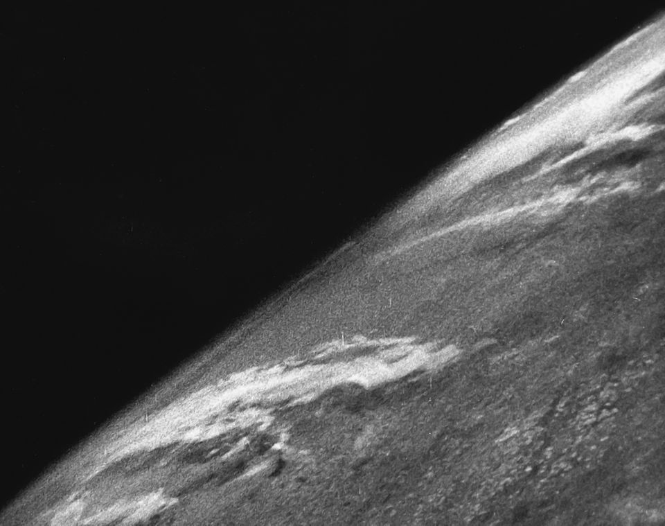 A black and white photograph of the earth from space, with the earth at a diagonal line across the frame. The left half is solid black and the right half is textured gray with light streaks.
