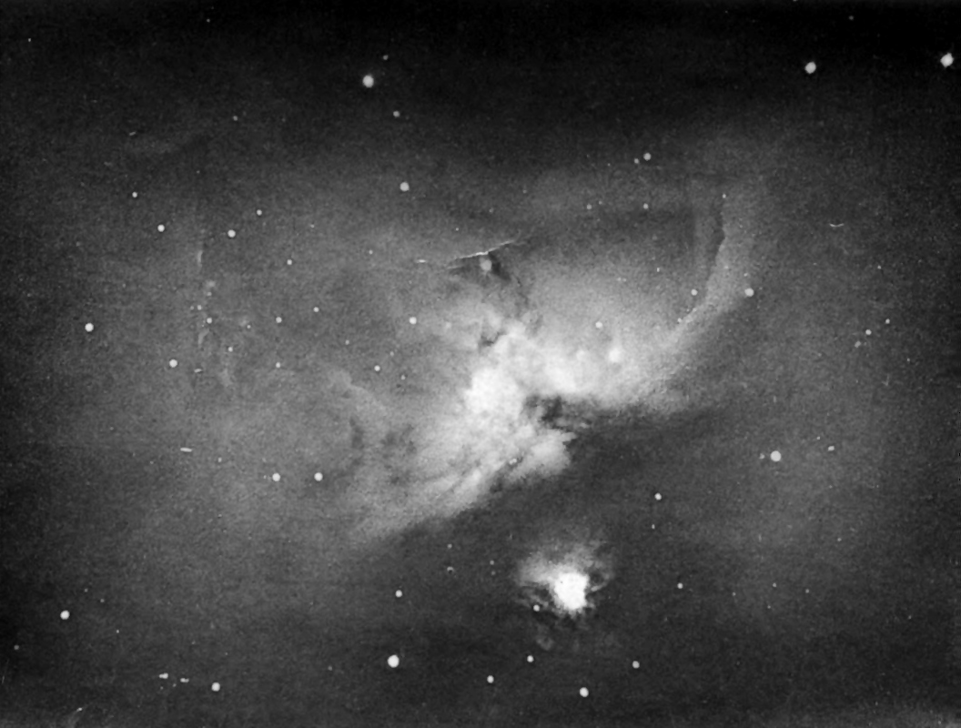 A black and white photograph of the Orion Nebula, with the lighter nebula more clearly visible and small white specs throughout the dark background.